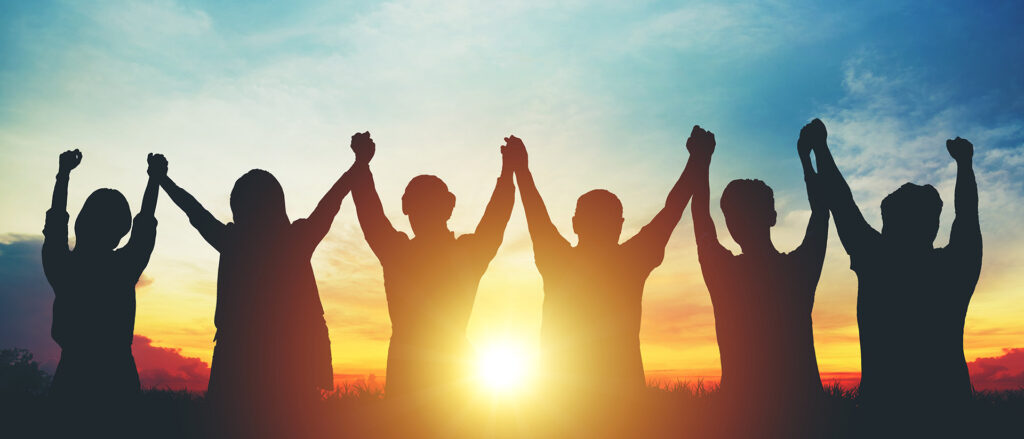 6 Menschen halten zusammen die Hände hoch und gucken in den Sonnenuntergang.